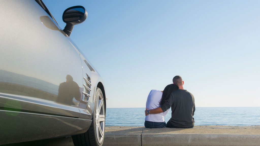 Illustrasjon av et par som sitter og ser utover sjøen med bilen parkert like ved.